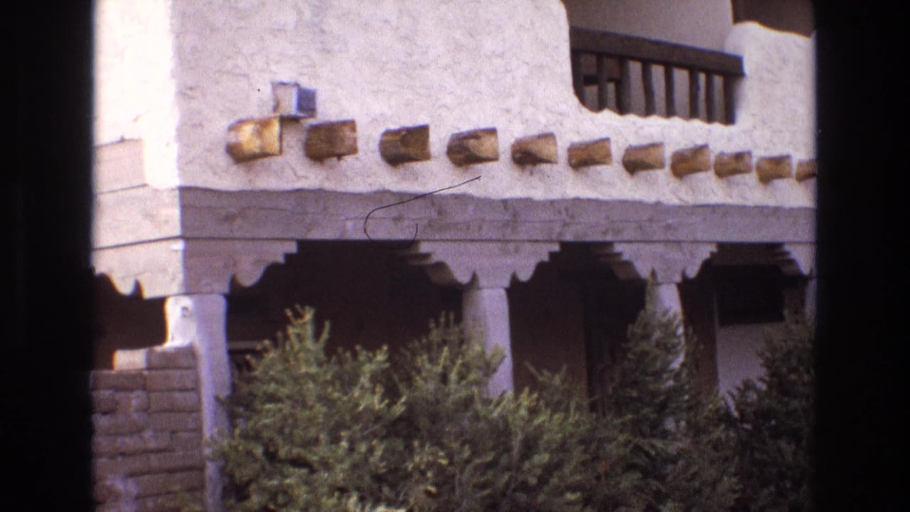 1973 New Mexico home-movie frame showing a white adobe-style building corner with projecting wooden vigas, shaded porch bays, support columns, and green shrubs in front.