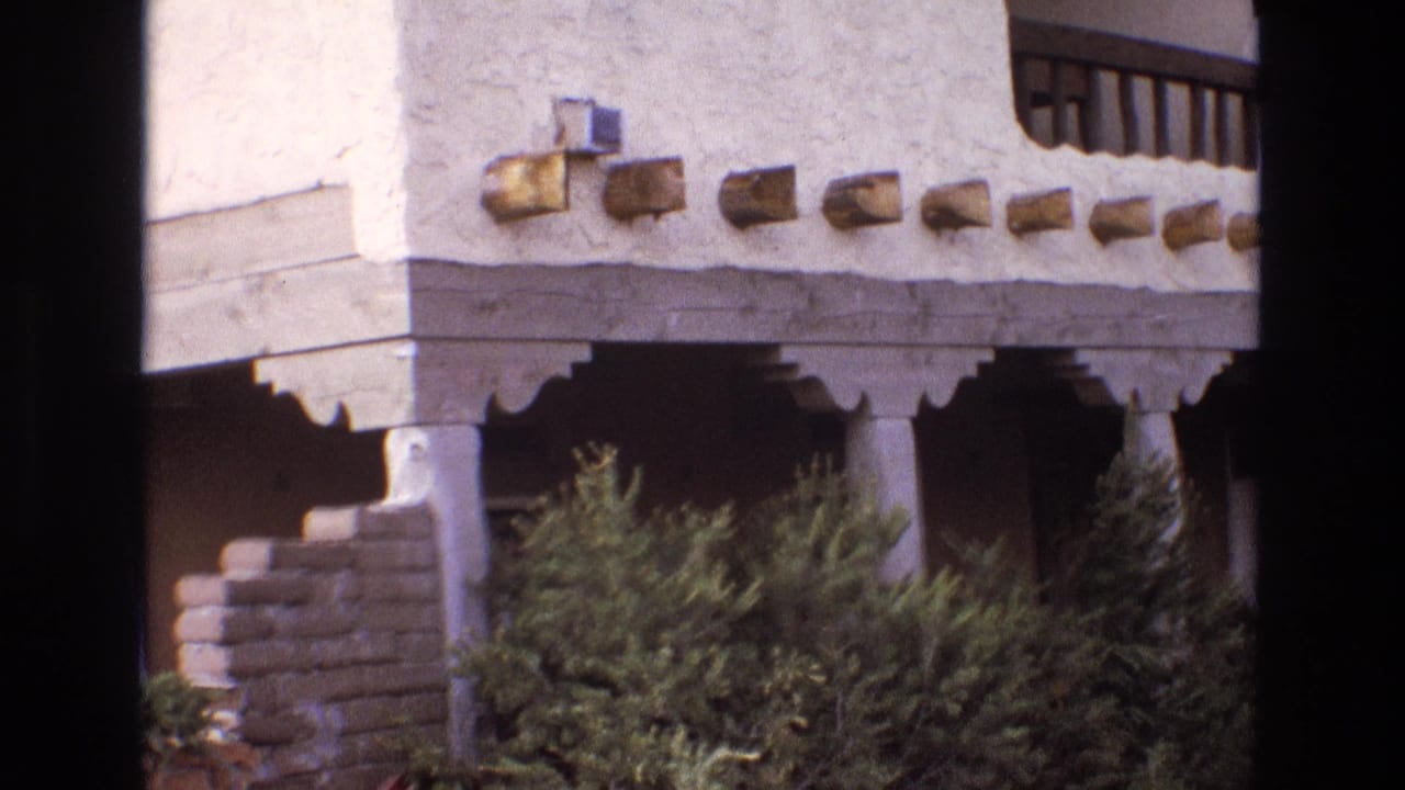 1973 New Mexico frame of an adobe-style facade with projecting wood beams, a shaded balcony or porch area, columns, shrubs, and dark vertical film-frame borders.