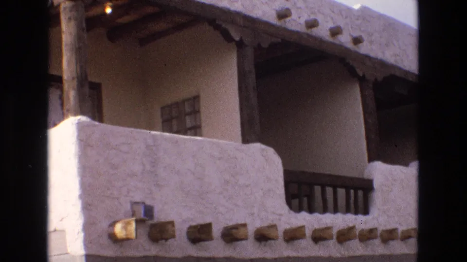 1973 New Mexico home-movie frame showing the upper exterior of a white adobe-style building with a balcony railing, projecting vigas, porch roof beams, and a small window.