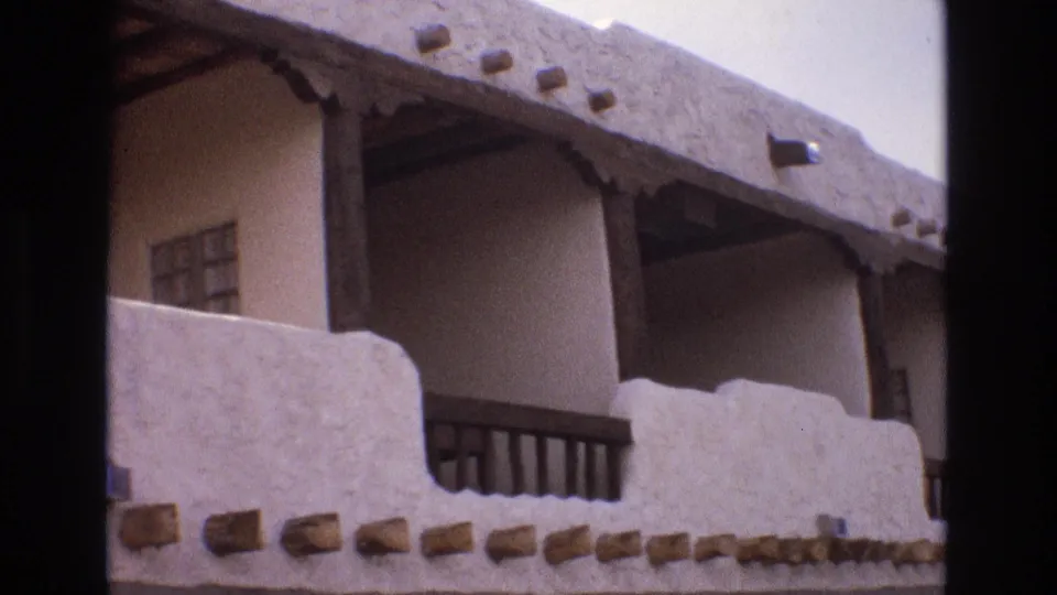 1973 New Mexico frame looking along a white adobe-style exterior with a dark balcony railing, exposed wooden vigas, shaded porch roof, and upper wall details.