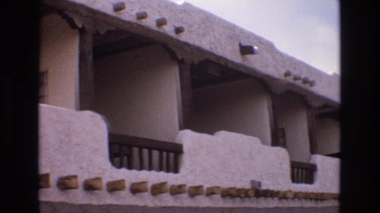 1973 New Mexico 8mm frame showing an adobe-style exterior with white stucco balcony walls, dark wood railings, projecting beams, and a pale sky above.