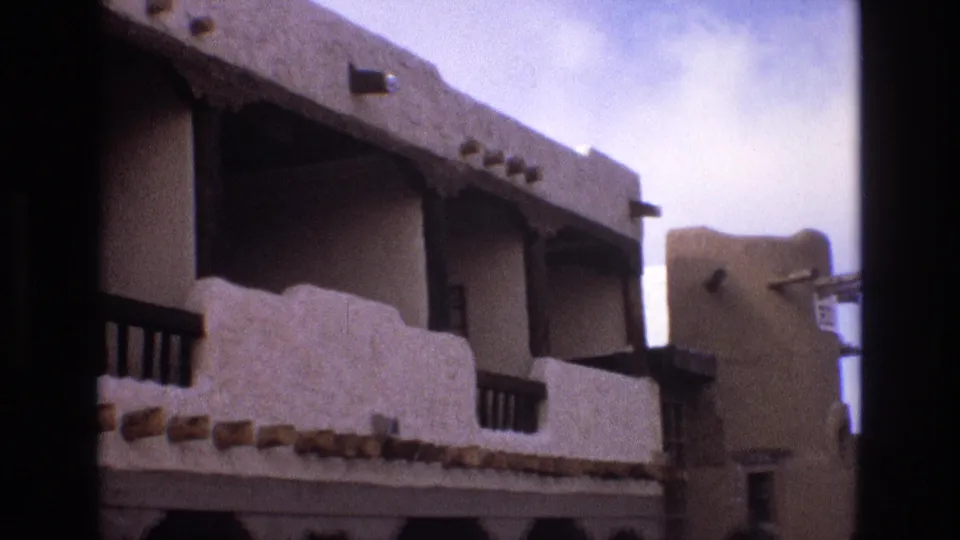 1973 New Mexico 8mm frame showing the camera panning along adobe-style balconies, with stucco walls, wood rails, and a neighboring earth-toned wall visible behind.
