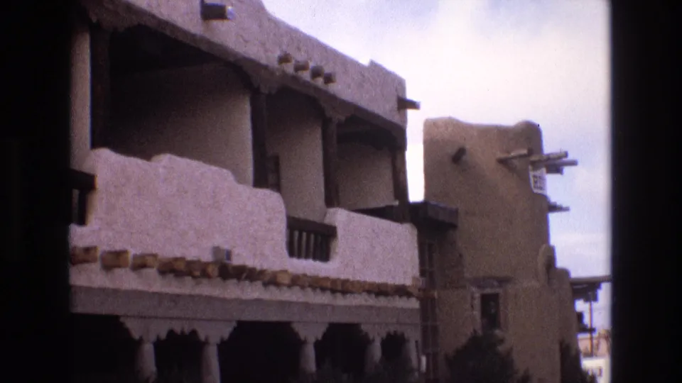 1973 New Mexico 8mm frame showing a stucco balcony facade with dark wood rails and projecting beams, plus an adjacent earth-toned building wall at right.