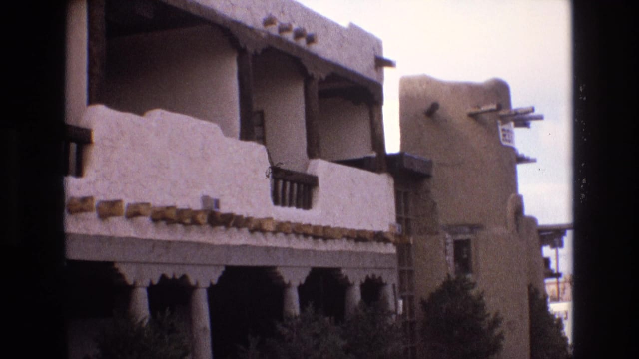 1973 New Mexico 8mm frame showing an adobe-style exterior with rounded stucco parapets, dark balcony railings, projecting beams, and shrubs along the lower level.
