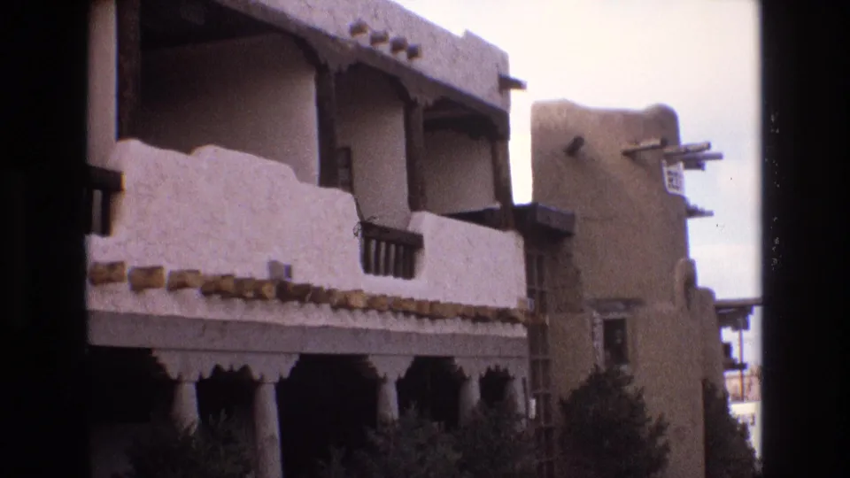1973 New Mexico 8mm frame showing an adobe-style exterior with rounded stucco parapets, dark balcony railings, projecting beams, and shrubs along the lower level.