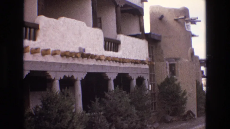 1973 New Mexico home-movie frame of a Southwestern-style building exterior with covered balcony, pale stucco walls, exposed beams, columns, and dense shrubs below.