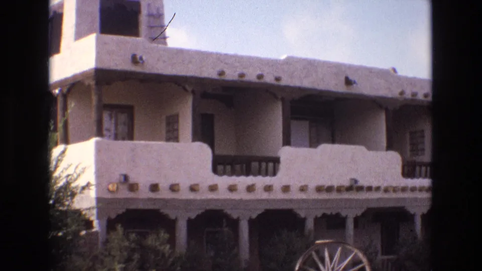 1973 New Mexico 8mm frame showing the upper facade of an adobe-style building with balcony railings, windows, exposed roof beams, decorative trim, and part of a wagon wheel at ground level.