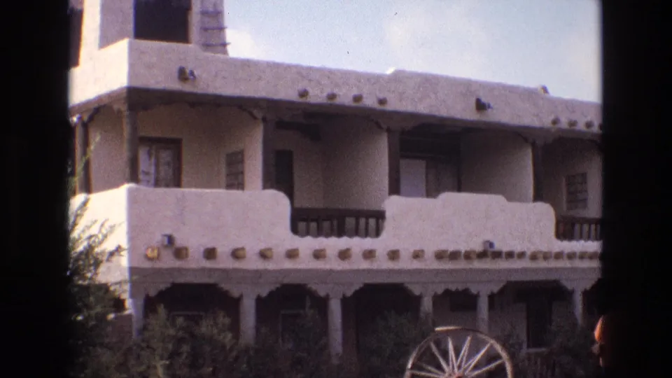 1973 New Mexico home-movie frame of an adobe-style exterior with a long balcony, pale stucco parapet, dark railings, lower arcade, shrubs, and a wagon wheel near the entrance area.