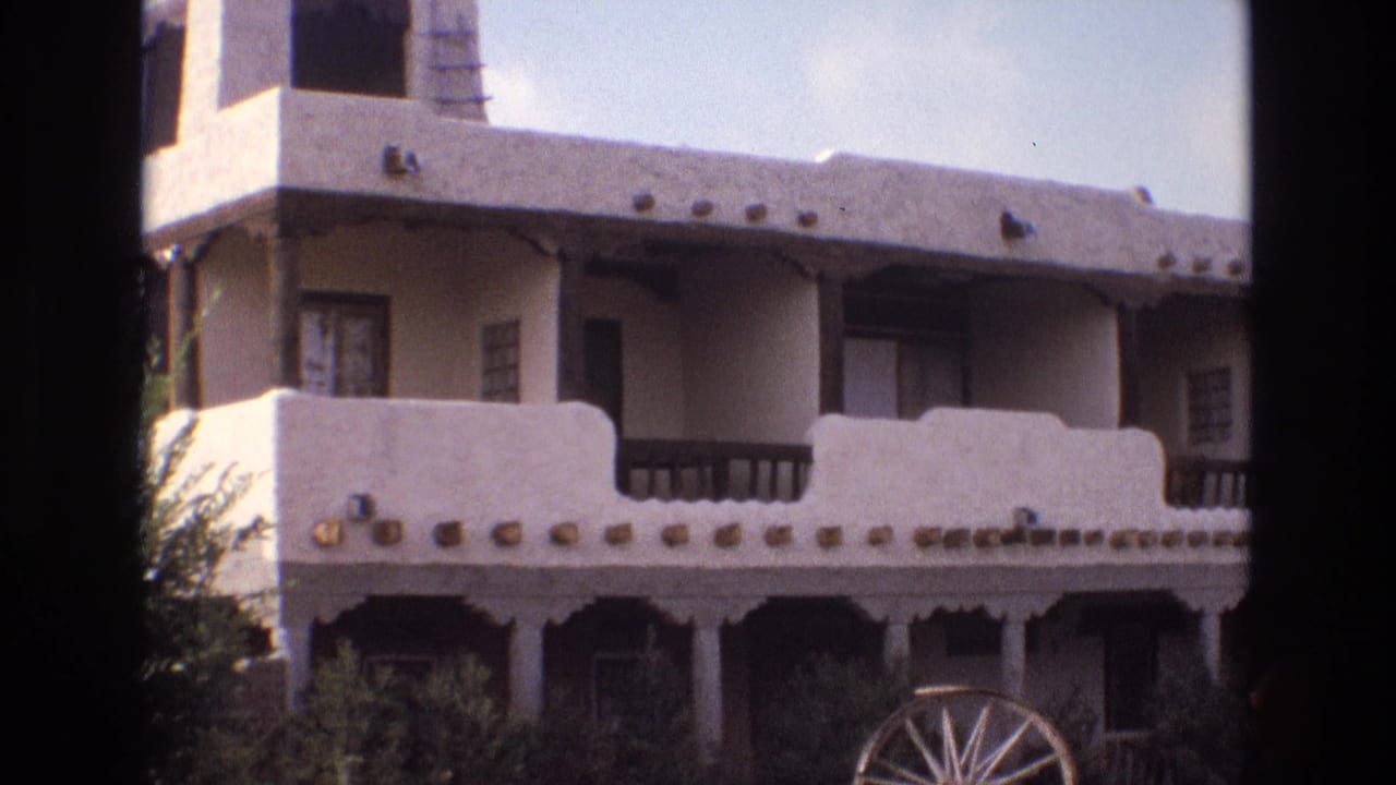 1973 New Mexico home-movie frame showing a white adobe-style stucco building with wooden balcony rails, decorative stone accents, shrubs, and a wagon wheel near the foreground.