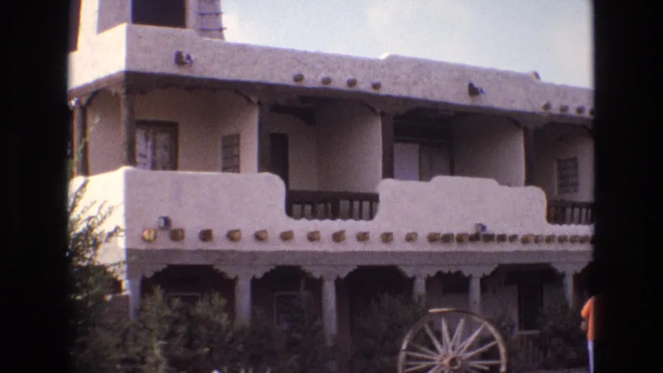 1973 New Mexico home-movie view of the same stucco building, with balcony details, a large wagon wheel, shrubs, and a partial adult figure near the right edge.