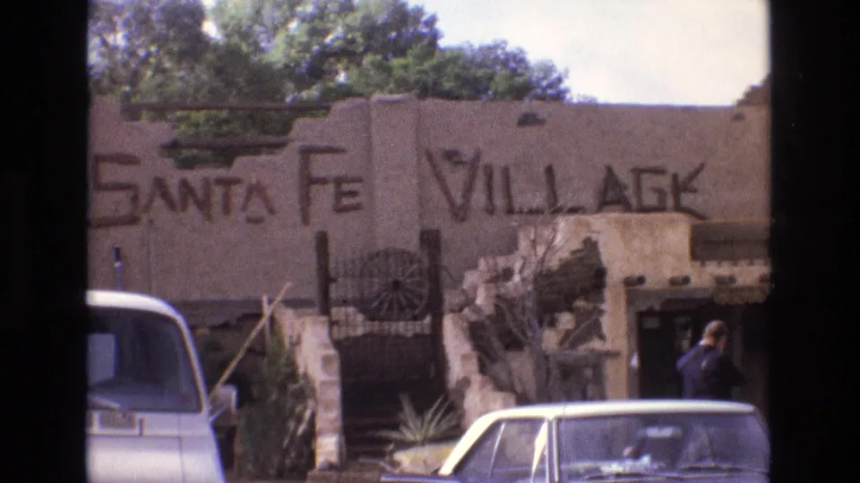 1973 New Mexico frame showing a wall painted with Santa Fe Village lettering, parked 1970s cars, wagon-wheel decoration, a stone entrance feature, and an adult walking at the right.