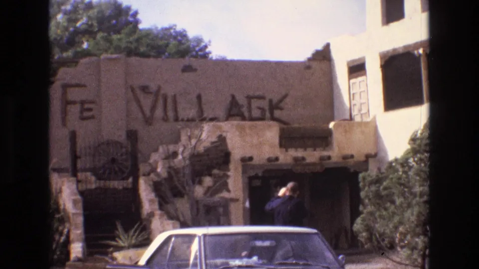 1973 New Mexico 8mm still showing the Santa Fe Village facade, dark entrance, adobe details, shrubs, and the roof of a parked white sedan.
