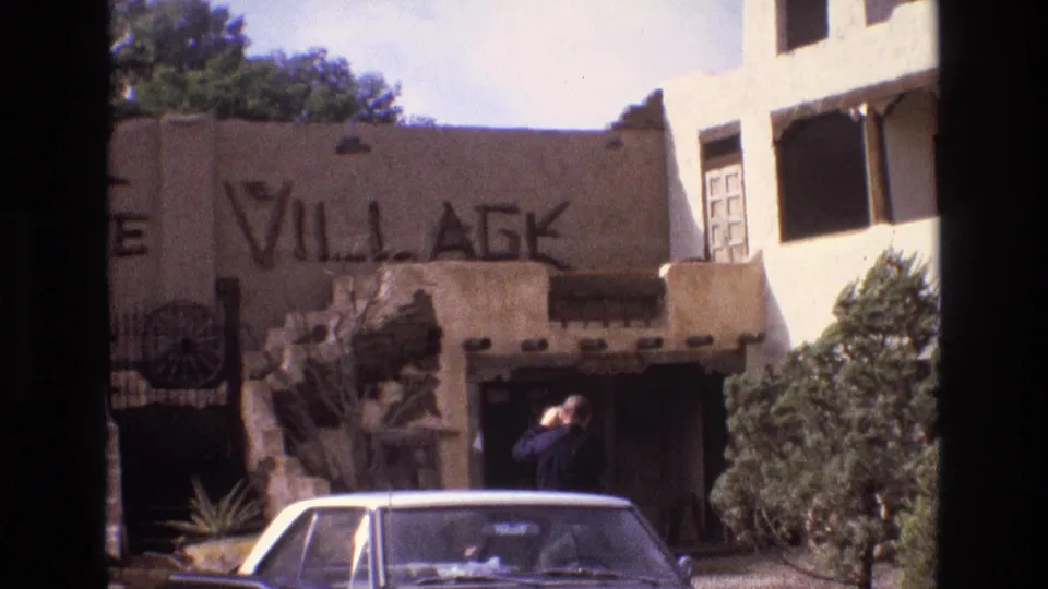 Final 1973 New Mexico frame showing an adult bending or turning near the Santa Fe Village entrance, with adobe-style walls, shrubs, and a parked white sedan in the foreground.