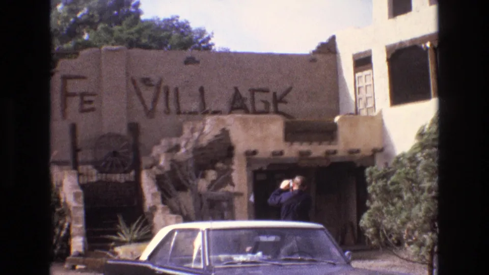 1973 New Mexico frame with a visitor holding a camera near the Santa Fe Village entrance while a classic sedan fills the lower foreground.