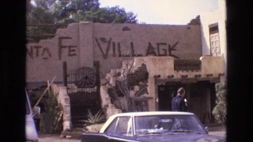 1973 New Mexico home-movie frame showing a visitor beside a parked sedan, with Santa Fe Village lettering, adobe-style architecture, desert planting, and a van at the edge of the frame.