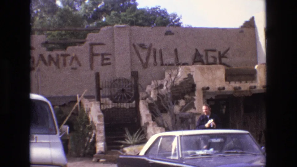 1973 New Mexico frame showing a man beside the foreground sedan with a vintage van partly visible, Santa Fe Village lettering, and rustic adobe-style exterior details behind him.