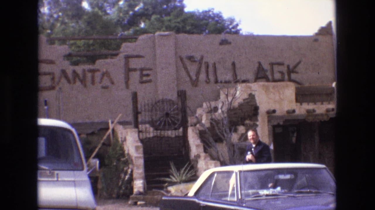1973 New Mexico home-movie frame showing an adult man outside a Santa Fe Village adobe-style facade, with parked classic cars and a wagon wheel gate visible.