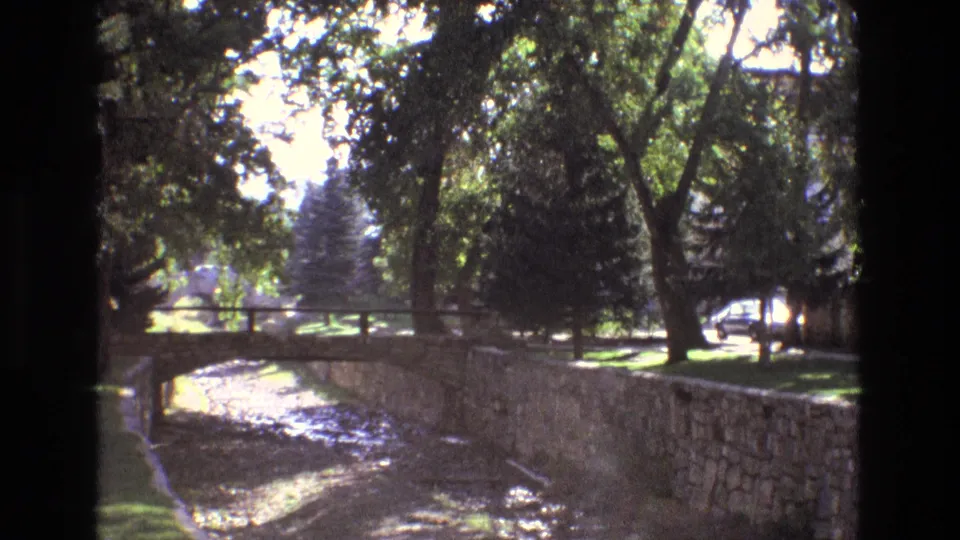 1973 New Mexico home-movie view of a dry creek or drainage channel bordered by a stone wall, trees, grass, and a small pedestrian or low bridge.