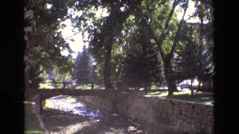 1973 New Mexico frame showing a shaded creekside area with a dry channel, stone retaining wall, small bridge, trees, lawn, and a parked car in the distance.