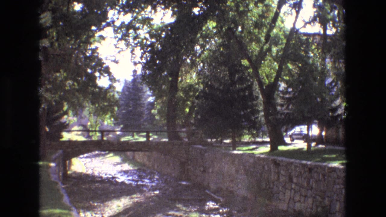 1973 New Mexico home-movie frame showing a stone-lined canal, small arched bridge, shaded trees, lawn, and a car passing on a background street.