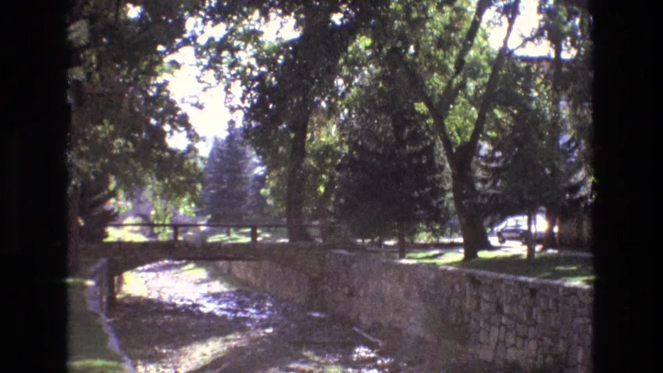 1973 New Mexico home-movie frame showing a stone-lined canal, small arched bridge, shaded trees, lawn, and a car passing on a background street.