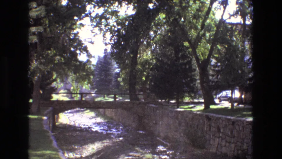 1973 New Mexico park view with a shallow canal, stone retaining wall, small bridge, dense tree canopy, and a light-colored car partly visible beyond the grass.