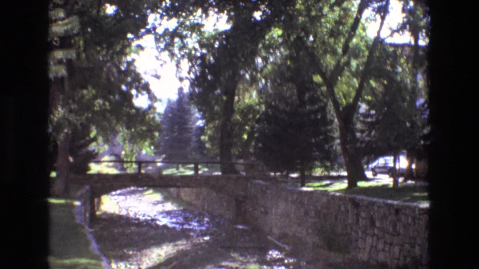 Soft 8mm frame of a New Mexico park canal in 1973, with a small bridge, stone wall, mature trees, dappled sunlight, and a street edge in the distance.