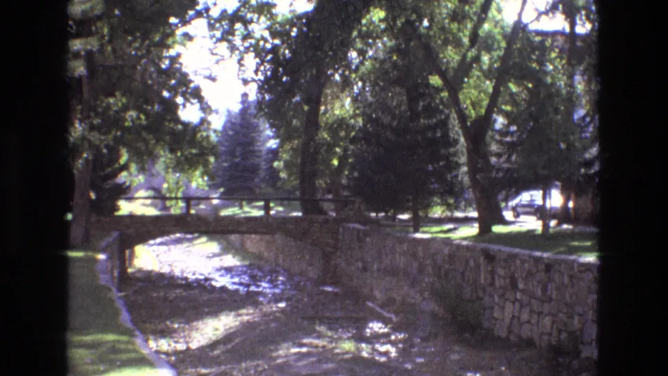 1973 New Mexico home-movie view of a curved stone-lined canal, a small park bridge, leafy trees, bright lawn, and a background roadway with no visible pedestrians.