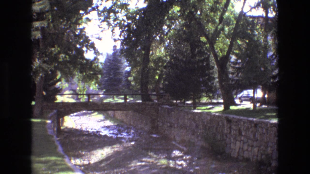 1973 New Mexico 8mm frame showing a stone-lined dry or shallow riverbed, small pedestrian bridge, grass, and leafy trees in bright daylight.