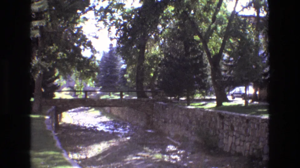 1973 New Mexico 8mm frame showing a stone-lined dry or shallow riverbed, small pedestrian bridge, grass, and leafy trees in bright daylight.