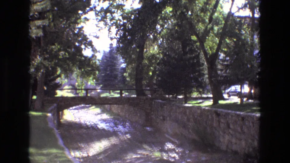 1973 New Mexico 8mm frame of the landscaped riverbed area, with stone walls, a small bridge, trees, grass, and strong sun-shadow contrast.