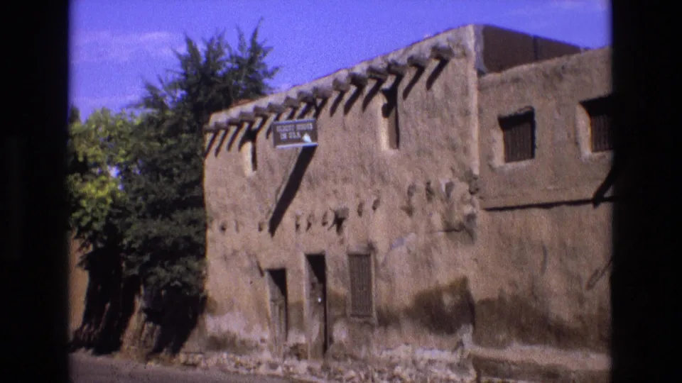 1973 New Mexico 8mm frame showing a historic adobe-style building exterior with projecting wooden beams, shuttered windows, trees, and a sign reading "Oldest House in USA."