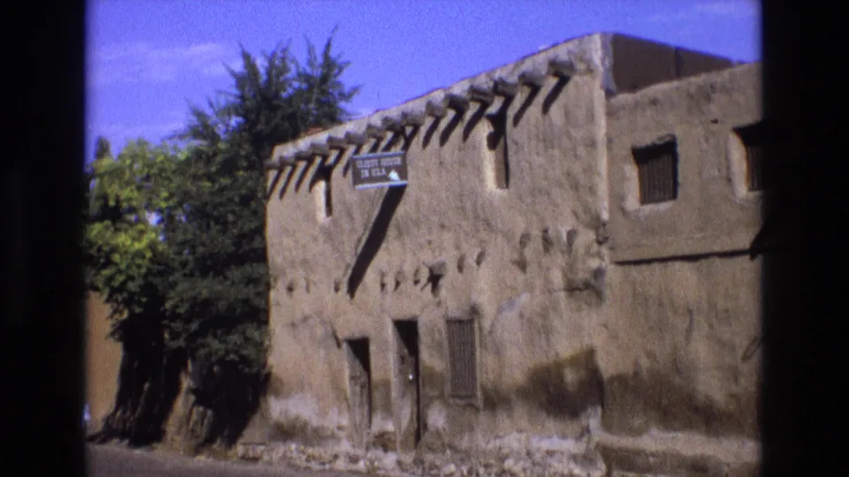 1973 New Mexico 8mm frame of the adobe landmark exterior, including the "Oldest House in USA" sign, wooden beams, small windows, nearby trees, and bright blue sky.