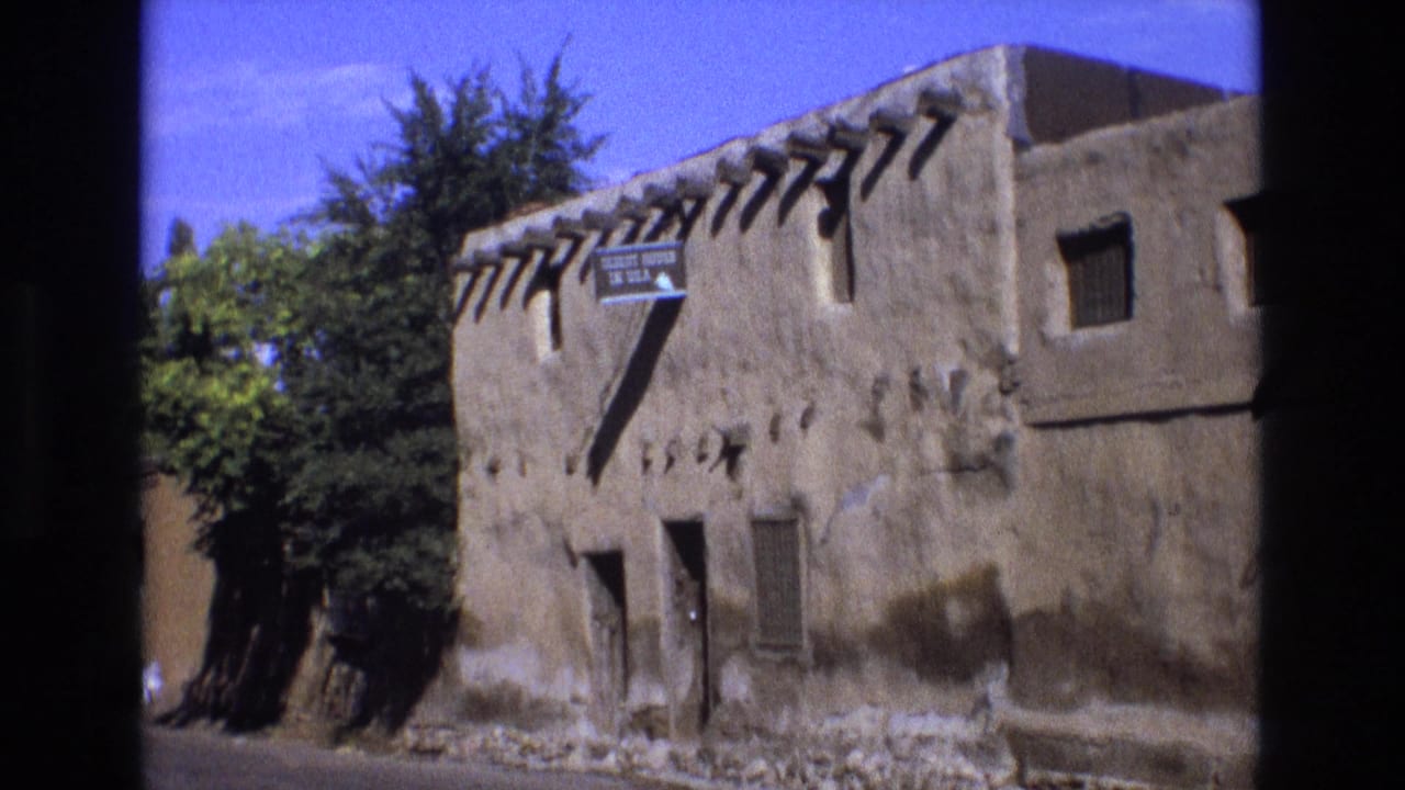 Sunlit adobe structure with wooden vigas, dark windows, trees, and readable Oldest House in U.S.A. sign.