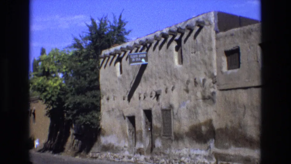 1973 New Mexico 8mm frame with a closer handheld composition of the adobe building, including projecting vigas, a tourist sign, small windows, trees, and sunlit earthen wall texture.