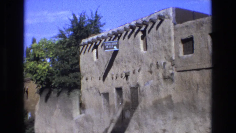 1973 New Mexico 8mm frame showing the adobe landmark exterior with a readable sign, projecting vigas, small windows, trees, bright sky, and handheld motion softness near the bottom of the frame.