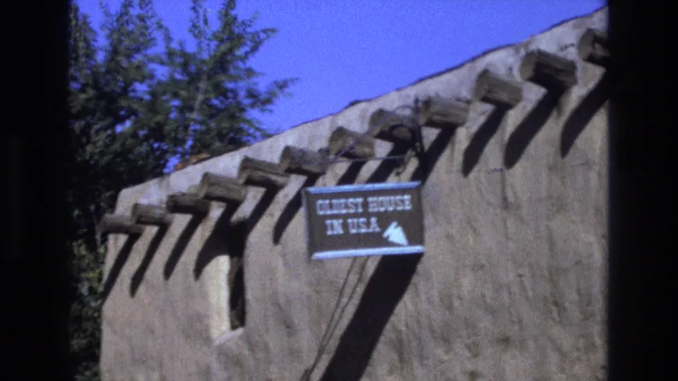 1973 New Mexico 8mm frame showing a tan adobe exterior with projecting wooden vigas, a blue directional sign reading “OLDEST HOUSE IN U.S.A.,” tree foliage, and bright sky.