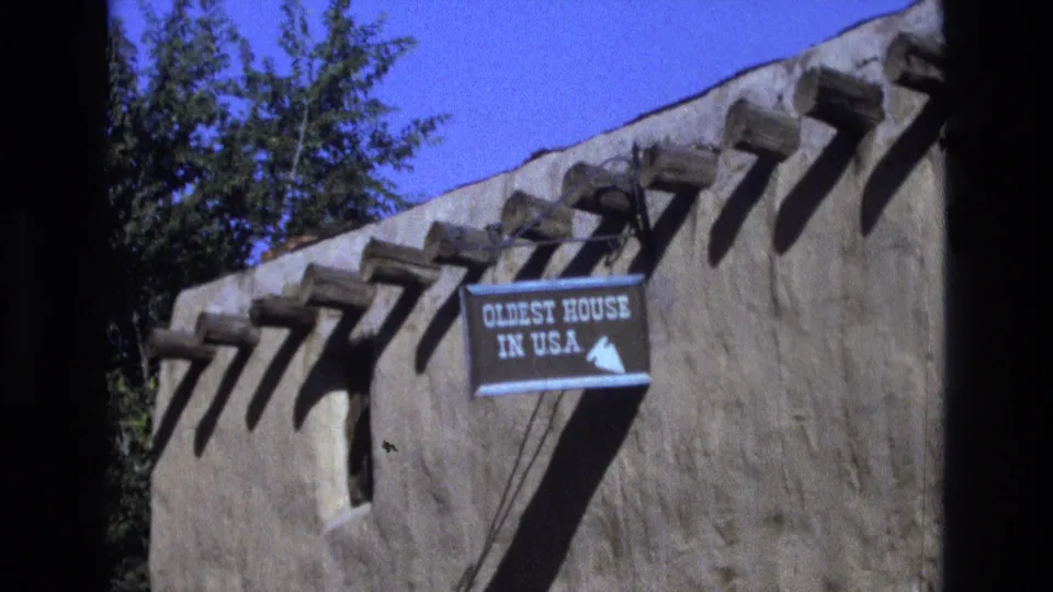 1973 New Mexico home-movie frame with a readable “OLDEST HOUSE IN U.S.A.” sign, arrow graphic, adobe wall surface, projecting roof vigas, and dark foreground film edge.