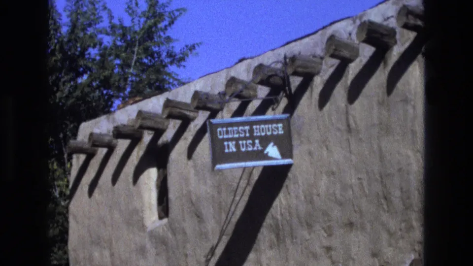 1973 New Mexico 8mm frame showing a closer handheld view of the adobe structure, wooden vigas casting shadows, and the blue “OLDEST HOUSE IN U.S.A.” sign.
