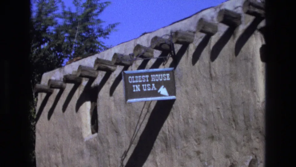 Final 1973 New Mexico frame of the adobe exterior with projecting wooden vigas, the “OLDEST HOUSE IN U.S.A.” directional sign, bright blue sky, and background foliage.