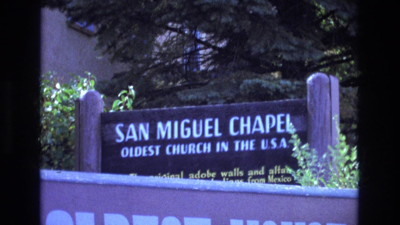 San Miguel Chapel sign partly framed by trees and adobe wall.