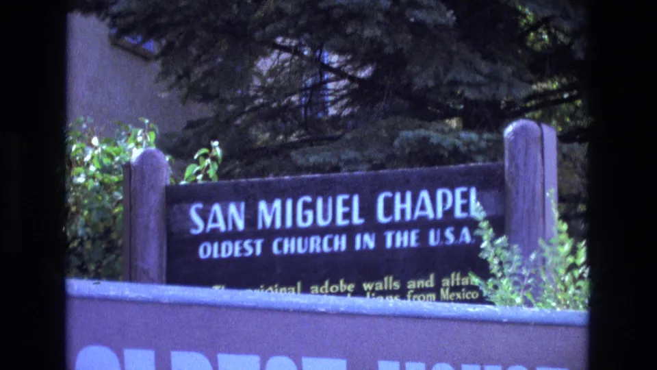 1973 New Mexico home-movie frame with a wooden San Miguel Chapel sign identifying it as the oldest church in the U.S.A., partly obscured by foreground wall and foliage.