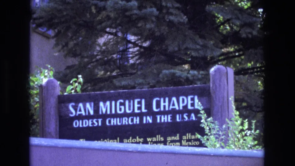 1973 New Mexico 8mm frame showing the San Miguel Chapel sign, its oldest church wording, wooden supports, and surrounding greenery.