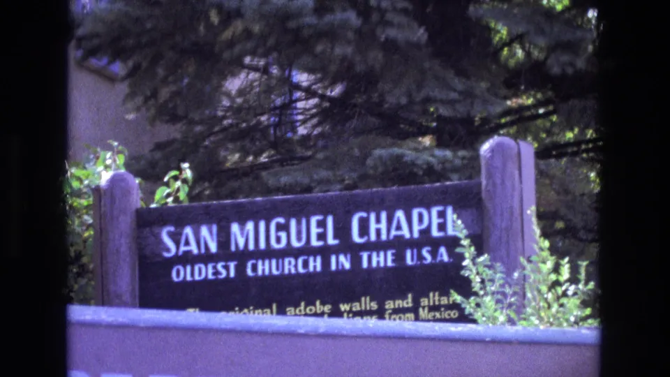 1973 New Mexico visitor-shot frame of the San Miguel Chapel sign across a low wall, with foliage and adobe-colored surfaces in the background.