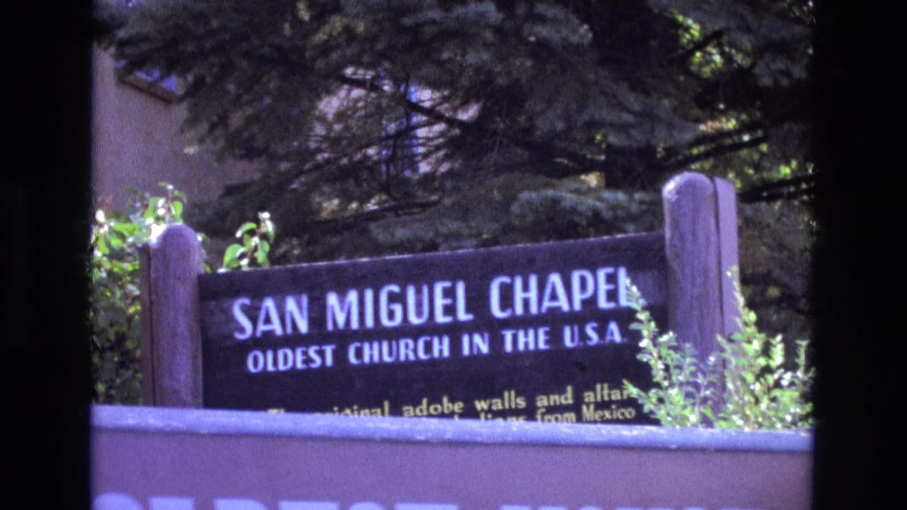 1973 8mm frame showing the San Miguel Chapel sign in New Mexico, with white lettering, wooden posts, greenery, and a low wall in front.