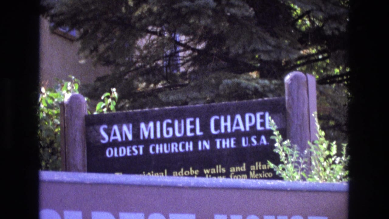 1973 New Mexico home-movie frame of the San Miguel Chapel sign, with the oldest church in the U.S.A. wording readable above shrubs and a foreground wall.