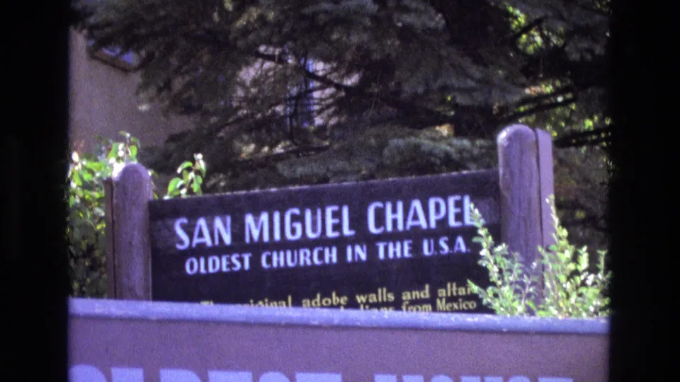 1973 New Mexico home-movie frame of the San Miguel Chapel sign, with the oldest church in the U.S.A. wording readable above shrubs and a foreground wall.