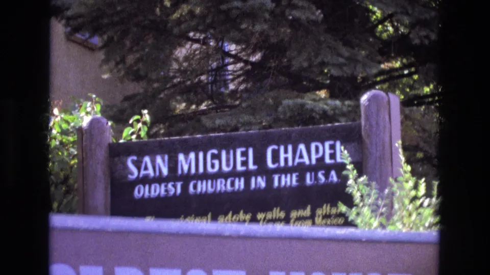 Soft 8mm frame from New Mexico showing the San Miguel Chapel sign at a slight angle, partly obscured by foliage and the low wall below.
