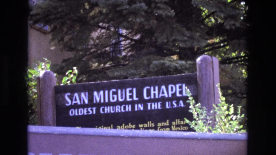 Final 1973 8mm frame of the San Miguel Chapel sign in New Mexico, with landmark lettering, shrubs, tree shade, and visible film-frame borders.