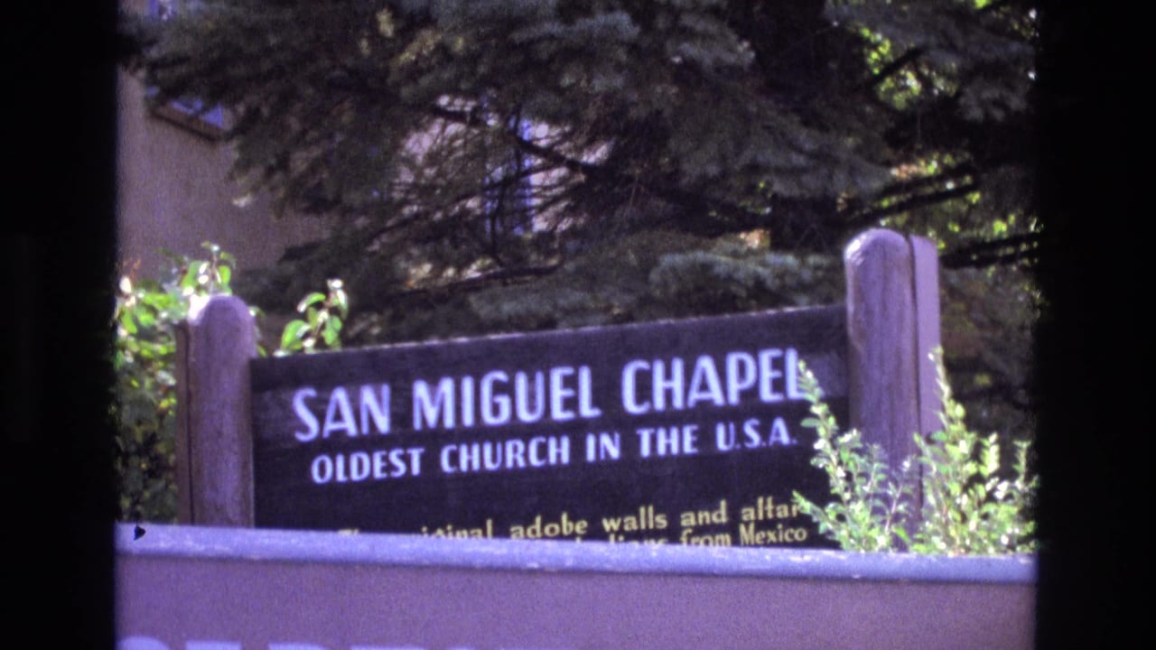 1973 New Mexico home-movie frame showing a sign for San Miguel Chapel beside trees, shrubs, and the edge of the chapel grounds.